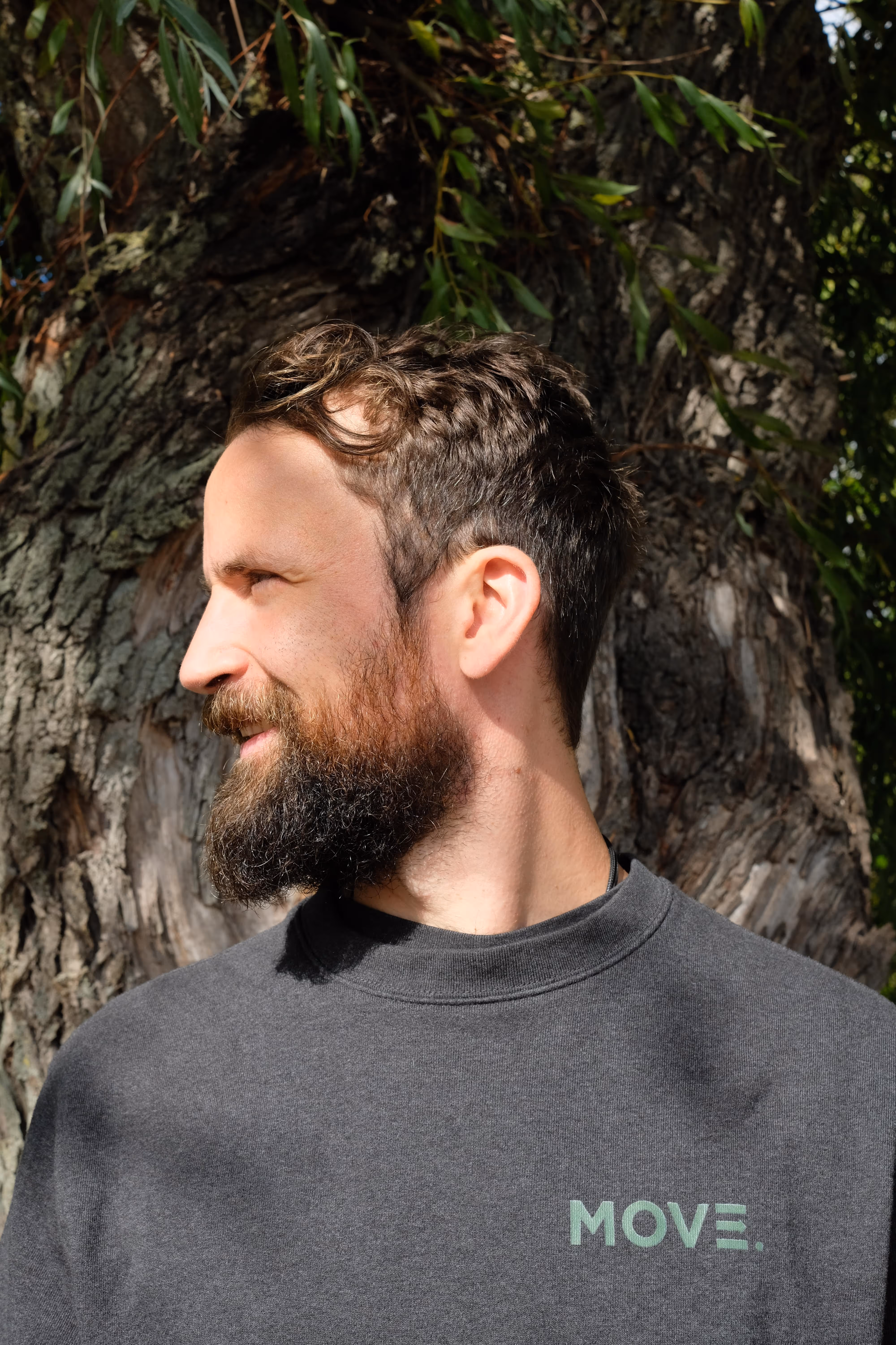 Rörelsecoach med skägg och mörkt hår som tittar åt vänster framför en stor trädstam.