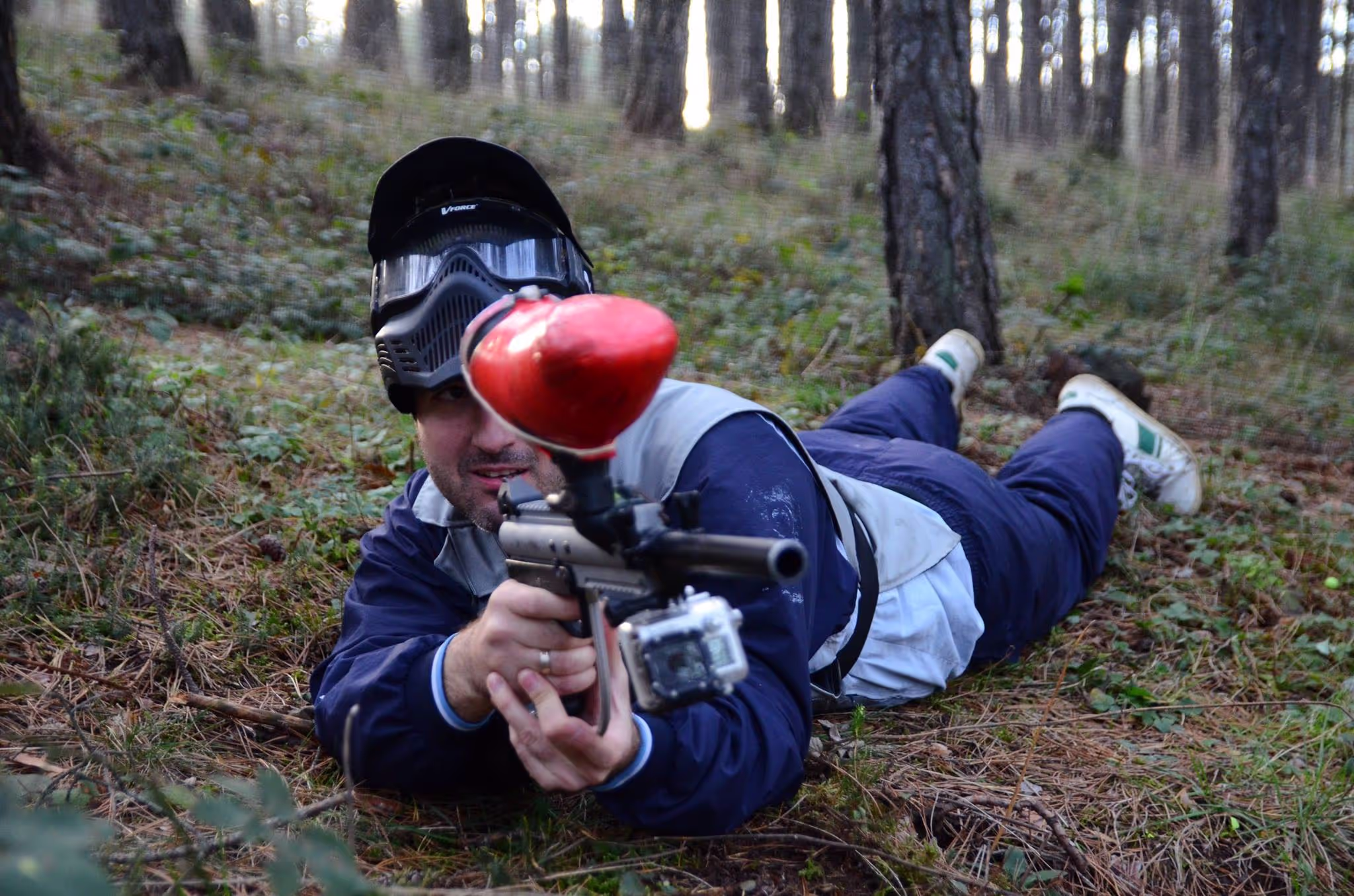 Čovek leži na zemlji u šumi i cilja metu sa paintball pištoljem, obučen u posebnu opremu za paintball.