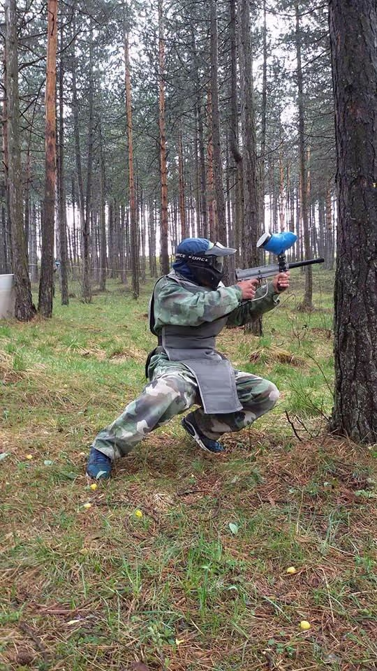 Čovek u opremi za paintball u toku igre, krije se iza stabala i gadja protivnika da bi pobedio.