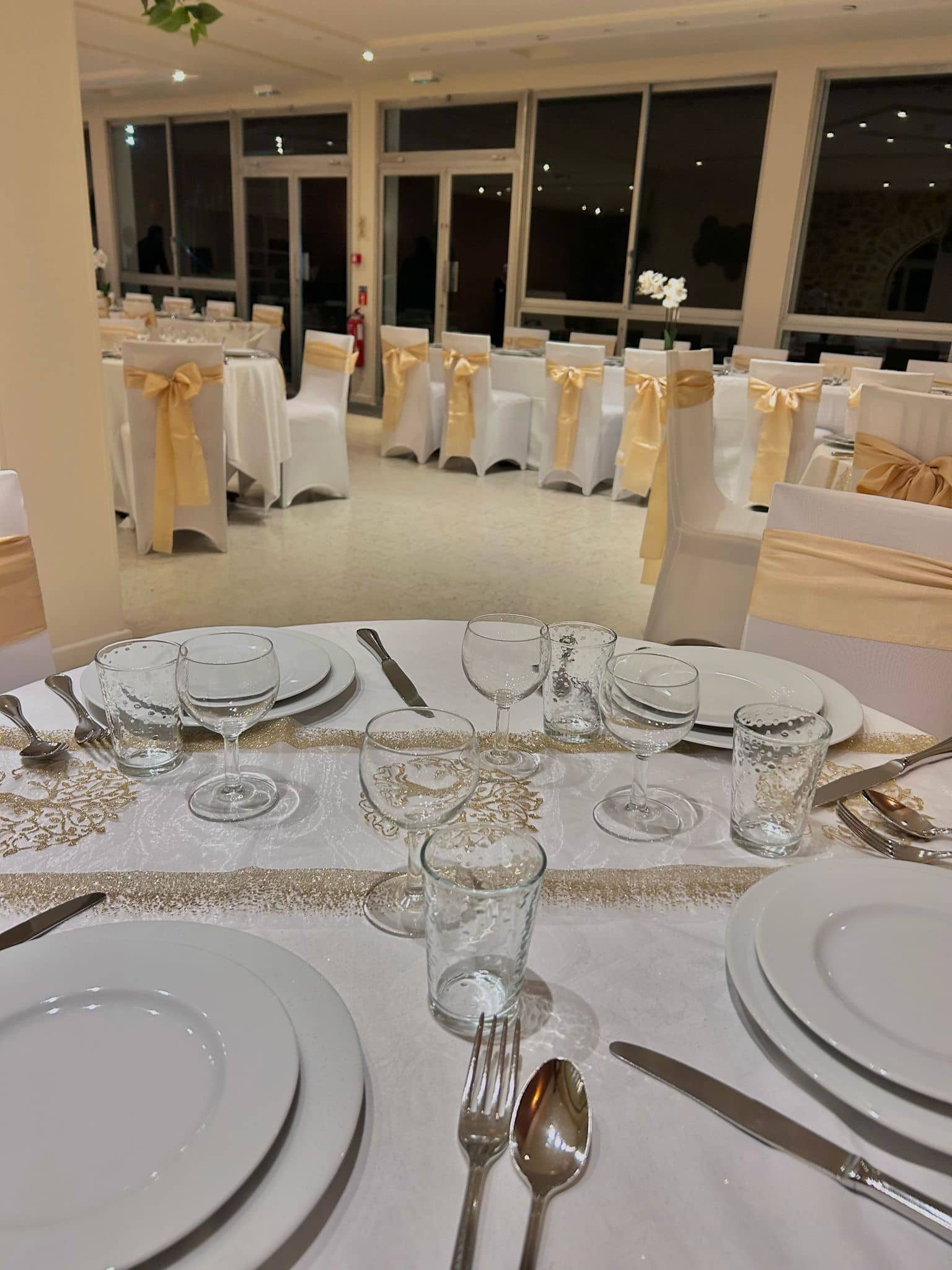 Table décorée pour un événement avec une nappe blanche ornée de motifs dorés, des verres à eau et à vin, ainsi que des assiettes et des couverts disposés soigneusement. Des chaises blanches avec des rubans jaunes sont visibles en arrière-plan, préparant l'espace pour un repas formel.