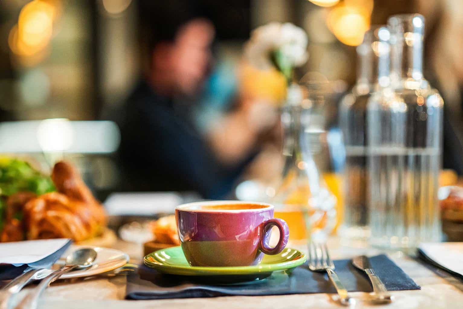 Une tasse de café colorée, avec une base violette et un fond vert, repose sur une soucoupe verte. La table est décorée avec des croissants et des ustensiles de cuisine. En arrière-plan, on aperçoit des bouteilles d'eau et des éléments flous d'une ambiance de café ou de brunch.