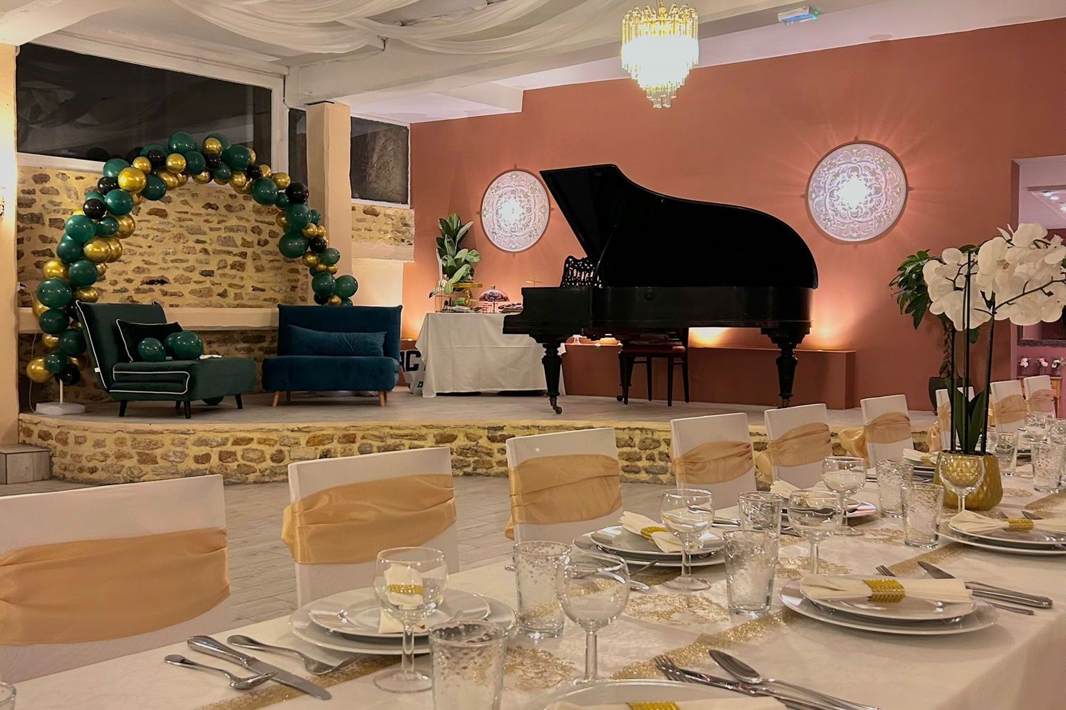 Une salle élégamment décorée pour un événement. En avant-plan, des tables dressées avec des assiettes blanches, des verres à vin et des serviettes pliées, avec des nappes blanches et un chemin de table doré. En arrière-plan, un canapé vert et une chaise assortie, décorés de ballons verts et dorés. Un grand piano noir est placé sur une estrade, accompagné de plantes et d'une table avec des desserts. Les murs sont décorés de lumières murales rondes, et le tout est éclairé dans des tons chaleureux.