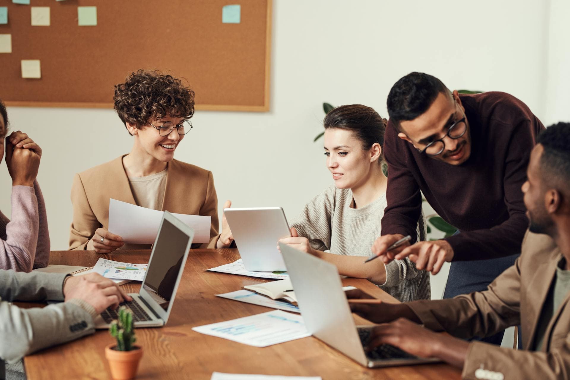Groupe de cinq personnes dans une réunion collaborative autour d'une table. Ils travaillent sur des ordinateurs portables, discutent et échangent des idées tout en analysant des documents imprimés et des tablettes.