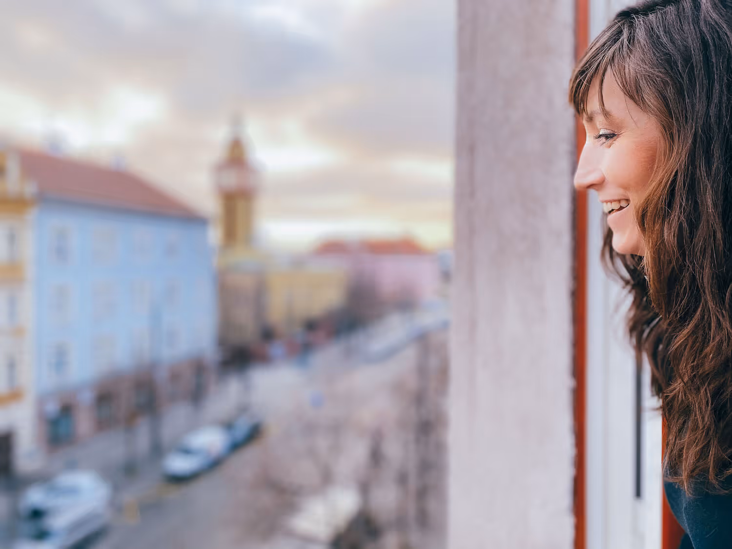 A profile of a young woman, looking out a window down to a street, smiling. 