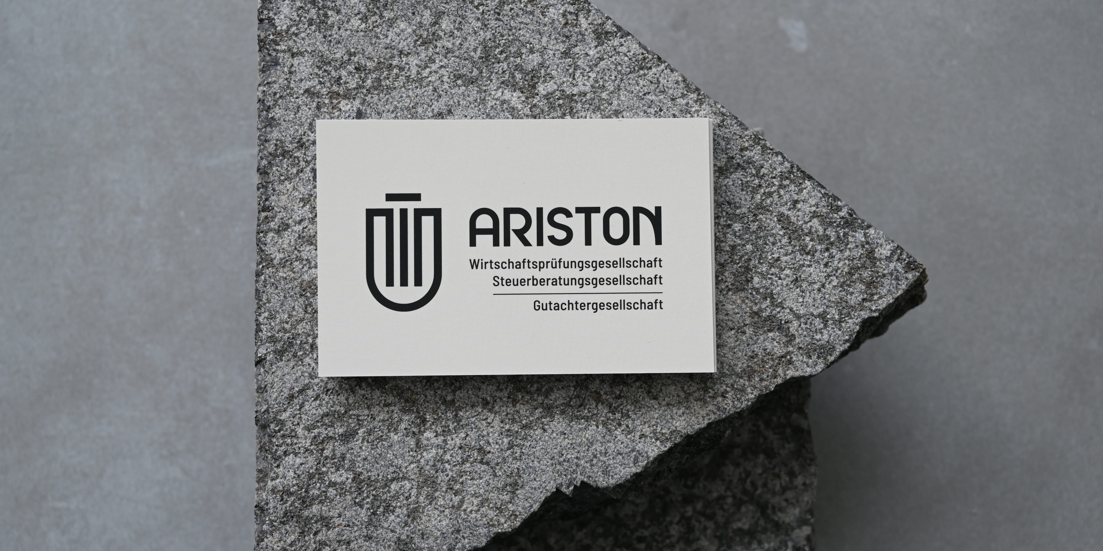 Visitenkarte mit ARISTON-Logo auf Granitplatte. Logo besteht aus stilisiertem Schildsymbol, daneben der Schriftzug „ARISTON“ mit den Begriffen „Wirtschaftsprüfung“, „Steuerberatung“ und „Gutachten“.