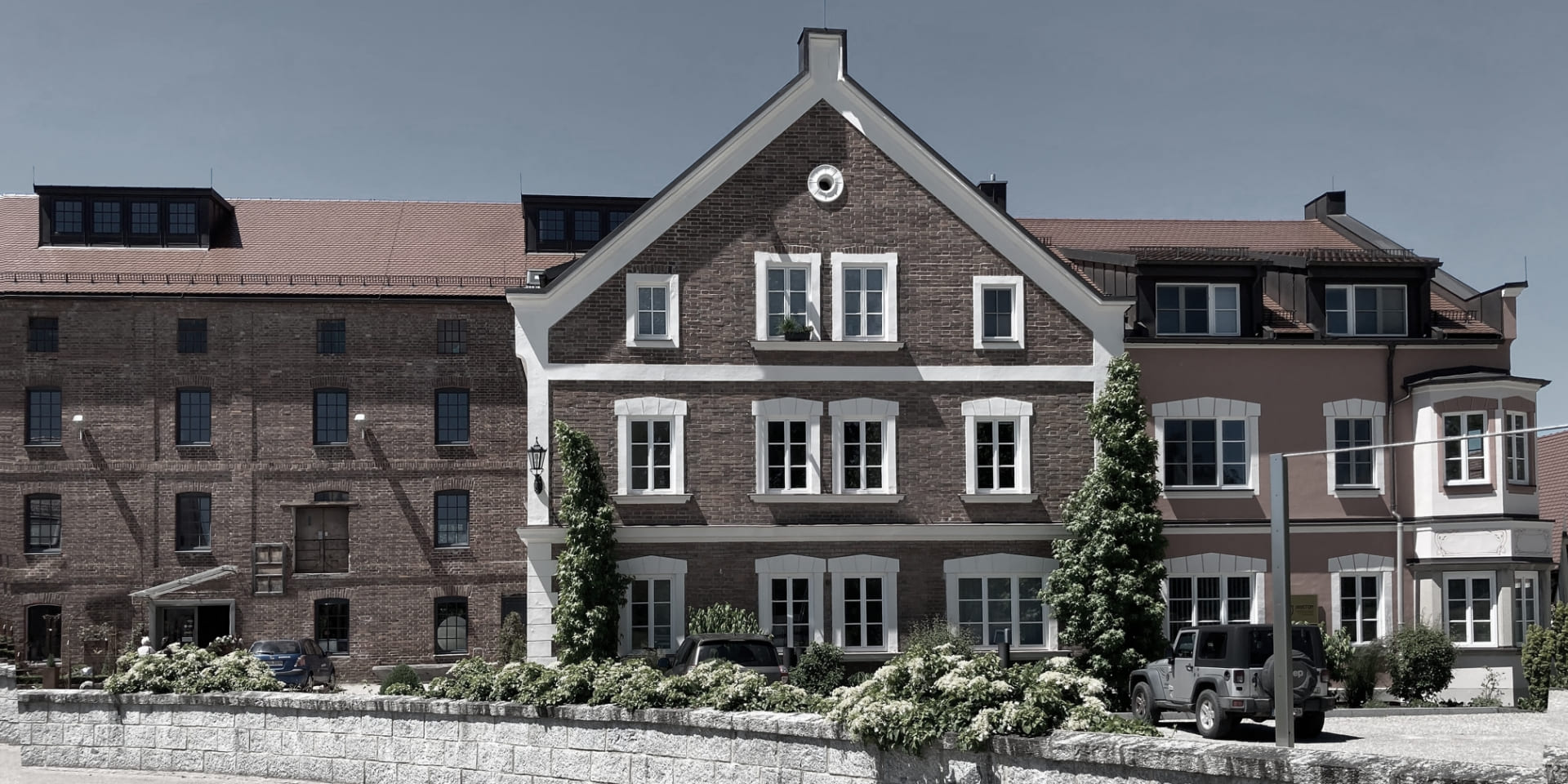 Bürogebäude mit Ziegelfassade, roten Dächern und weissen Fensterrahmen. Im Vordergrund Blumenbeet, Mauer und mehrere parkierte Autos.