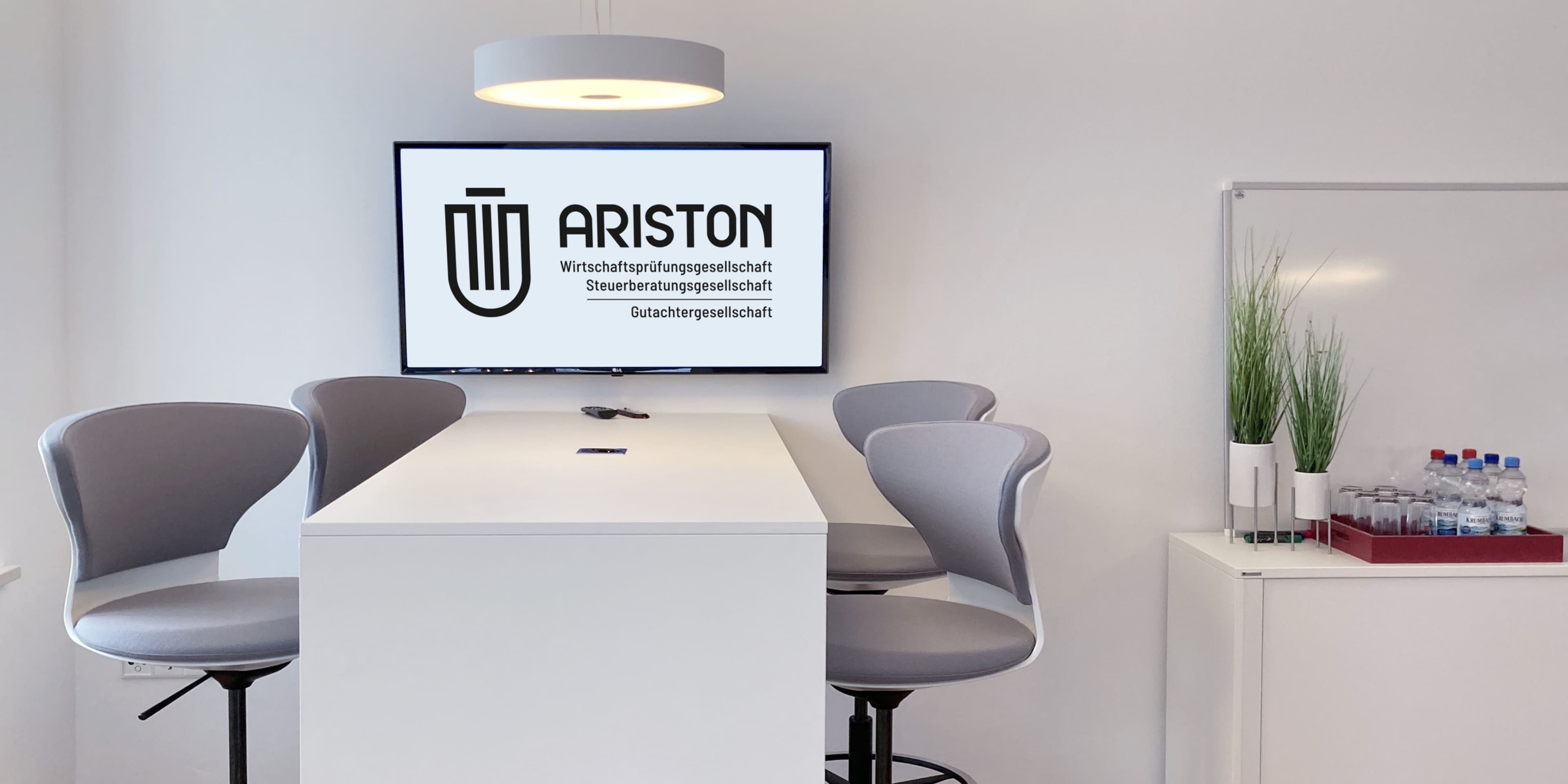 Besprechungsraum mit hellem Tisch und vier grauen Barhockern, im Hintergrund ein Monitor mit ARISTON-Logo und Getränke auf einem Sideboard.