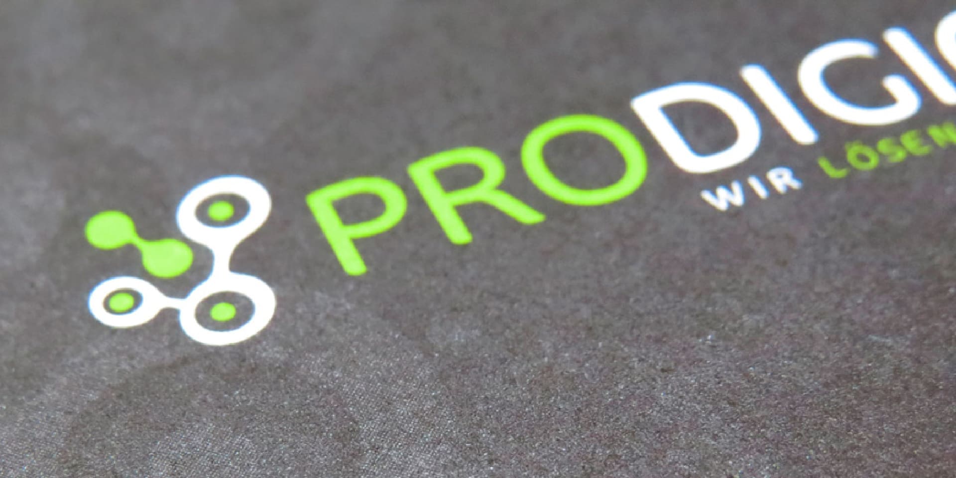 Detailaufnahme des PRODIGIOO-Logos mit grünem und weissem Netzwerk-Symbol auf schwarzem Papier. Prodigioo Wasserzeichen im Hintergrund.