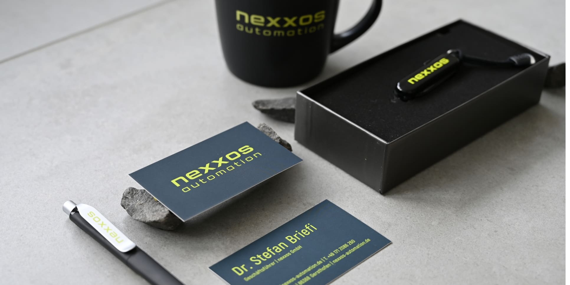 Visitenkarten, Kugelschreiber, USB-Stick und Tasse mit „nexxos automation“-Branding auf grauem Untergrund, modernes Corporate Design in Grün und Dunkelblau.