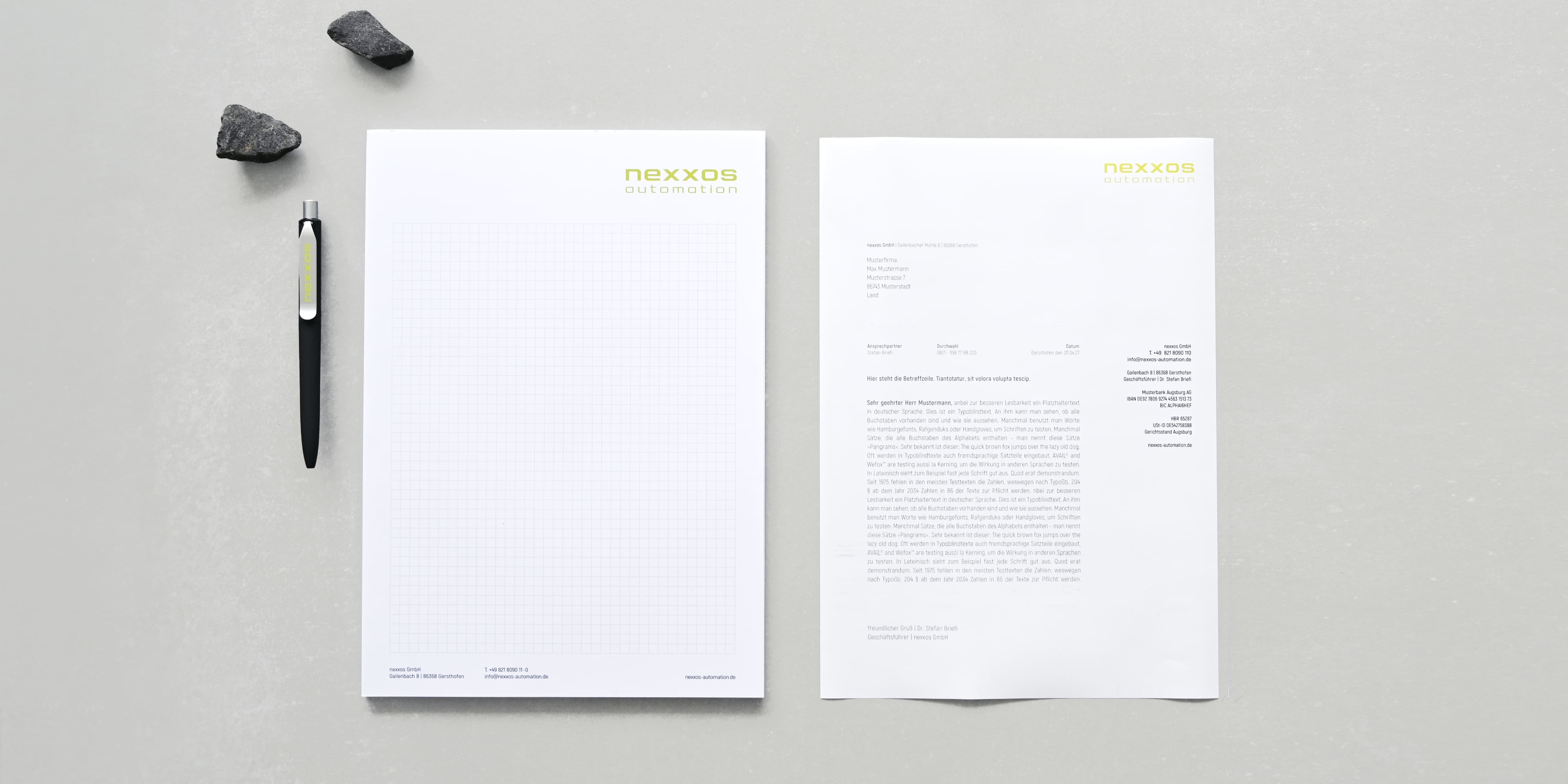 Briefpapier-Set von nexxos automation bestehend aus kariertem Notizblock, vorgedrucktem Brief und Kugelschreiber, arrangiert auf hellgrauer Oberfläche.