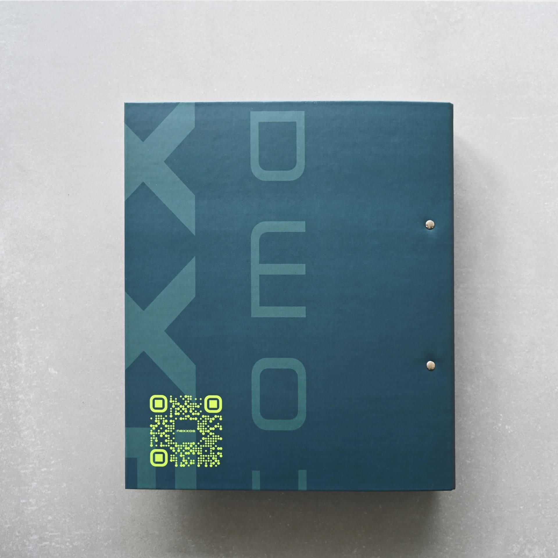 Dunkelblauer Ordner mit senkrechtem Schriftzug „nexxos“ und QR-Code in Neongelb auf hellem Untergrund fotografiert.