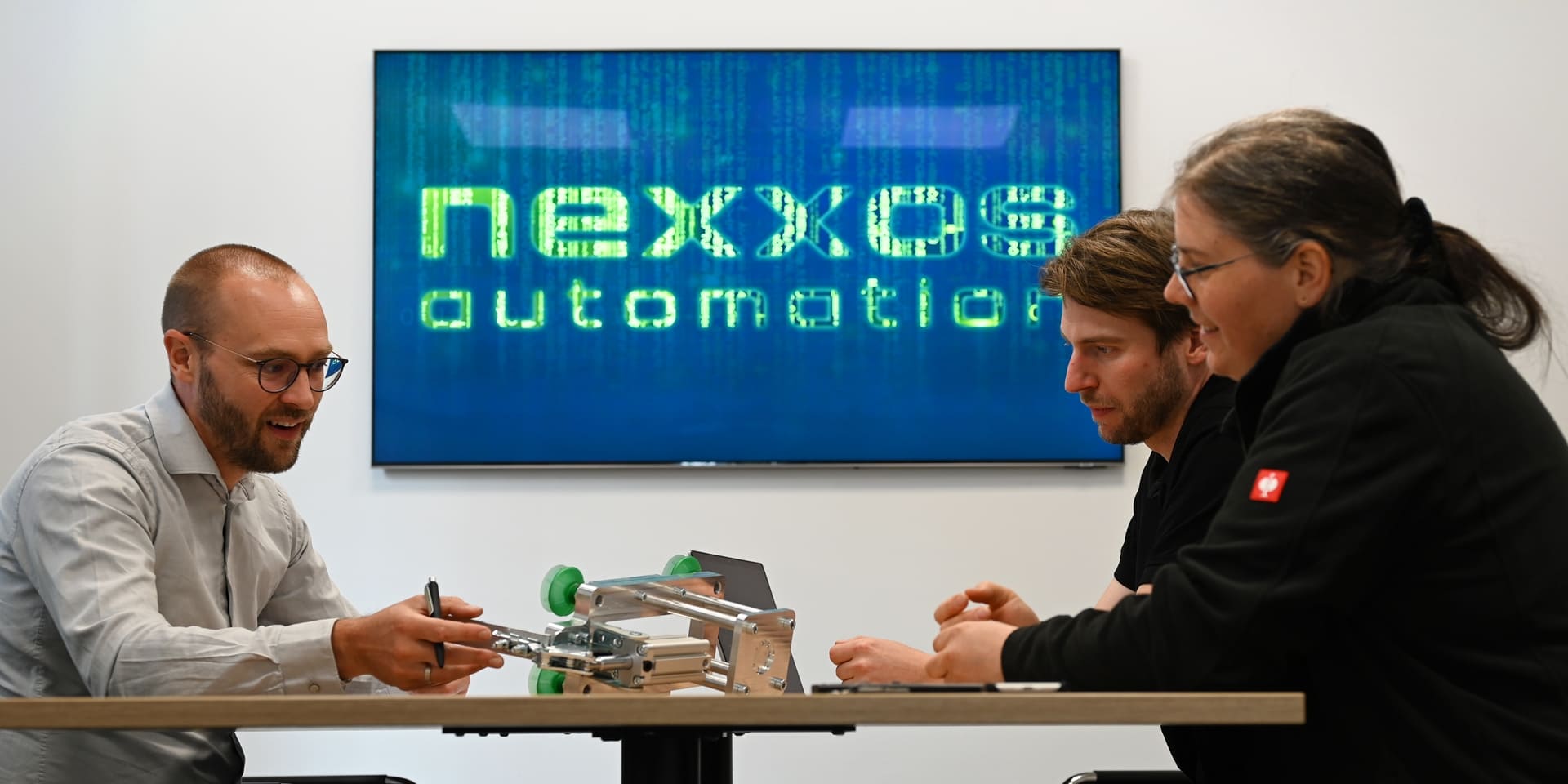 Drei Personen im Meetingraum diskutieren über ein Bauteil, im Hintergrund ein Bildschirm mit dem leuchtenden Logo von nexxos automation.