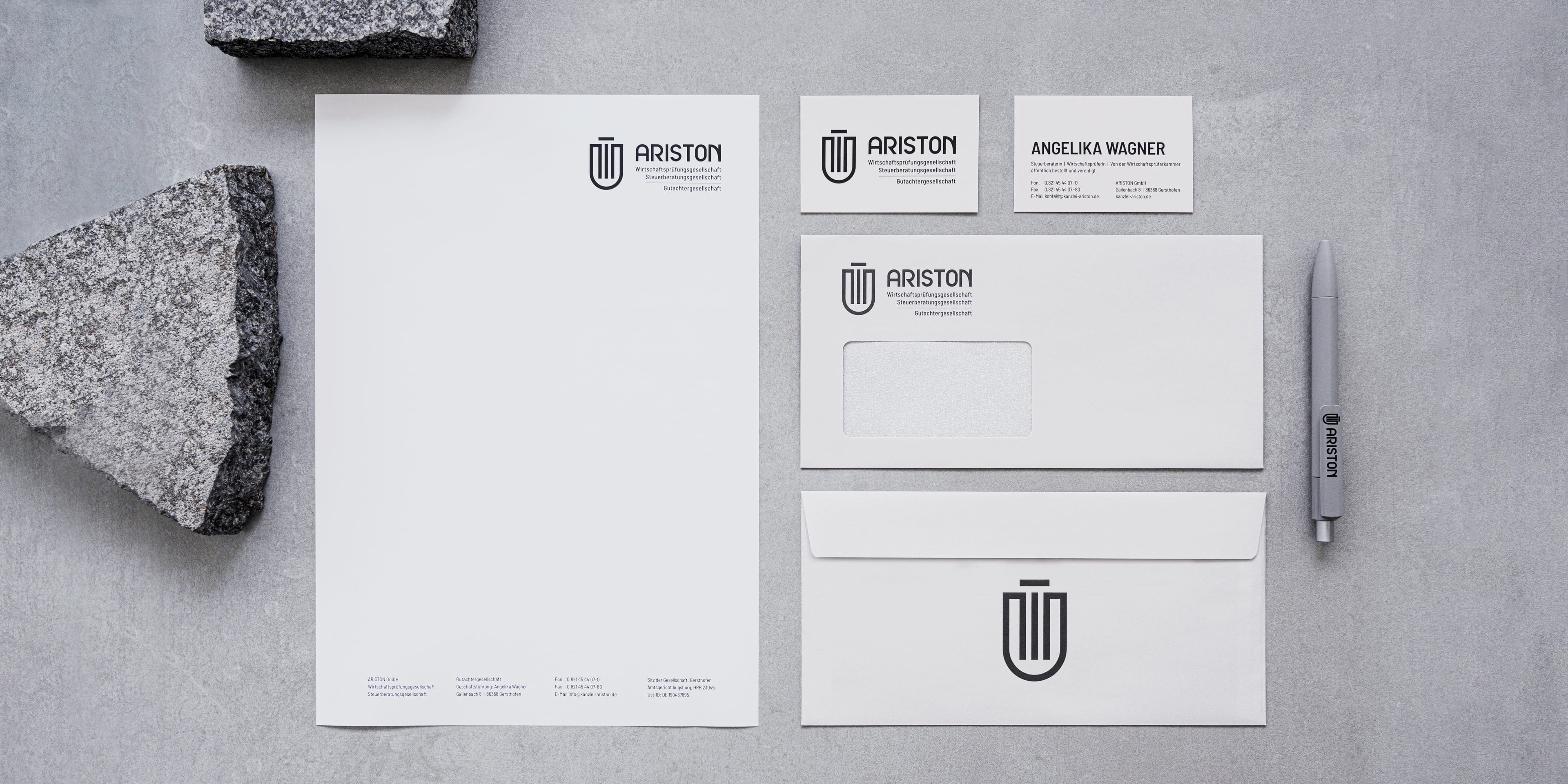 Geschäftsausstattung von ARISTON auf grauem Untergrund mit Steinfragmenten. Zu sehen sind puristisch gestaltetes Briefpapier, Visitenkarten, Umschläge und ein Kugelschreiber mit Logo.