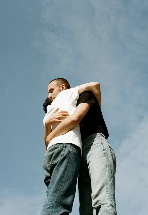 Two men embracing each other outdoors against a blue sky with clouds.