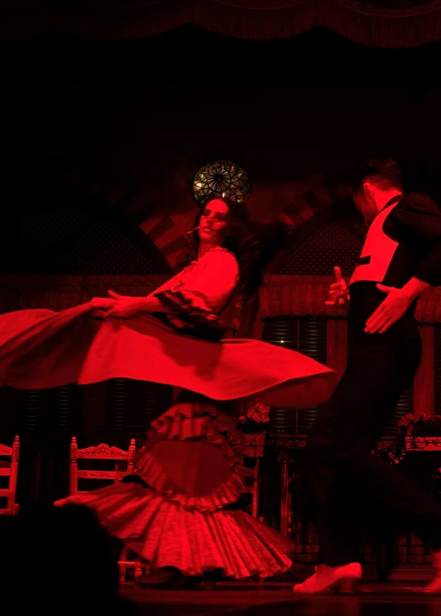 Two dancers performing flamenco on stage under red lighting, the female dancer twirling her flowing skirt.