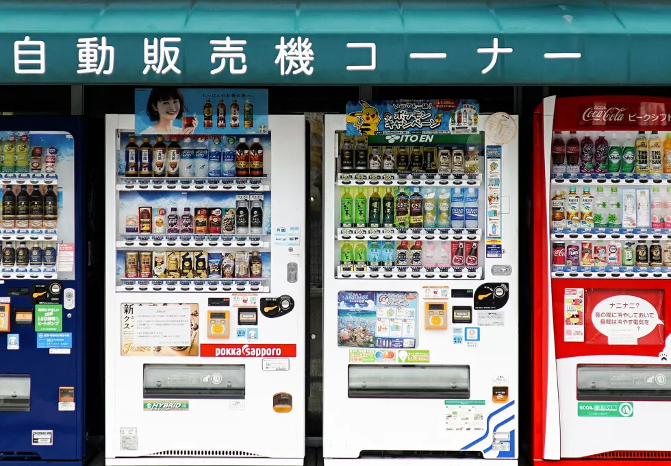 Three Japanese vending machines side by side stocked with various beverages including tea, coffee, soft drinks, and water under a green awning with Japanese text.