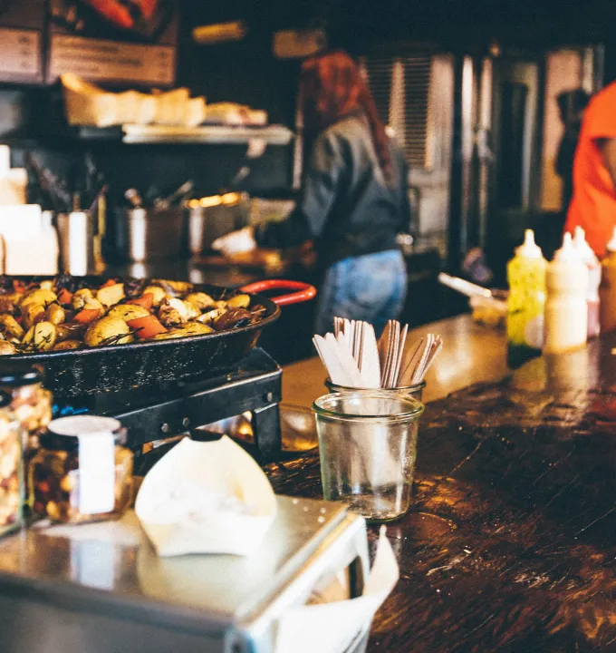 Large pan of cooked roasted potatoes and vegetables on a counter with utensils in a glass jar and a person working in the background.