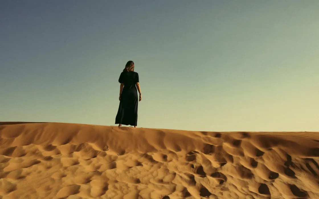 A person in a long black dress standing alone on a sandy desert dune under a clear sky.