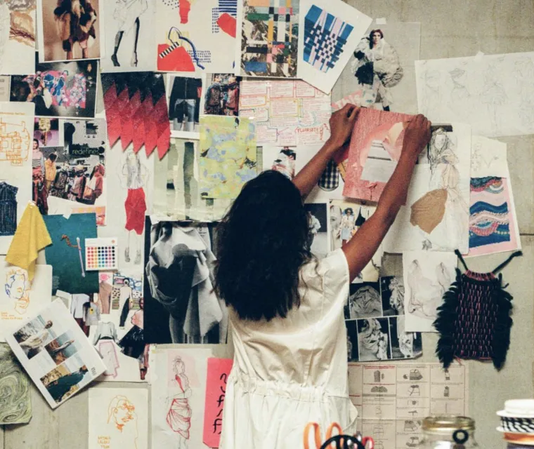 Person with dark hair in a white dress arranging artwork and sketches on a wall filled with fashion designs and colorful patterns.