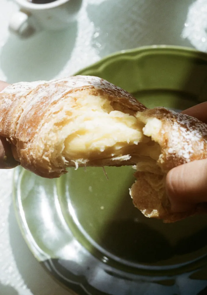 Hands tearing apart a powdered sugar-covered croissant with creamy filling over a green plate.