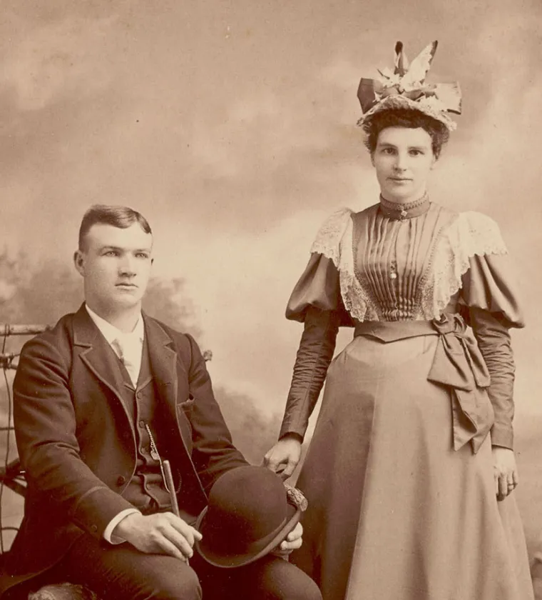 Sepia-toned vintage portrait of a man seated holding a bowler hat and a woman standing beside him wearing a long dress and a decorative hat.