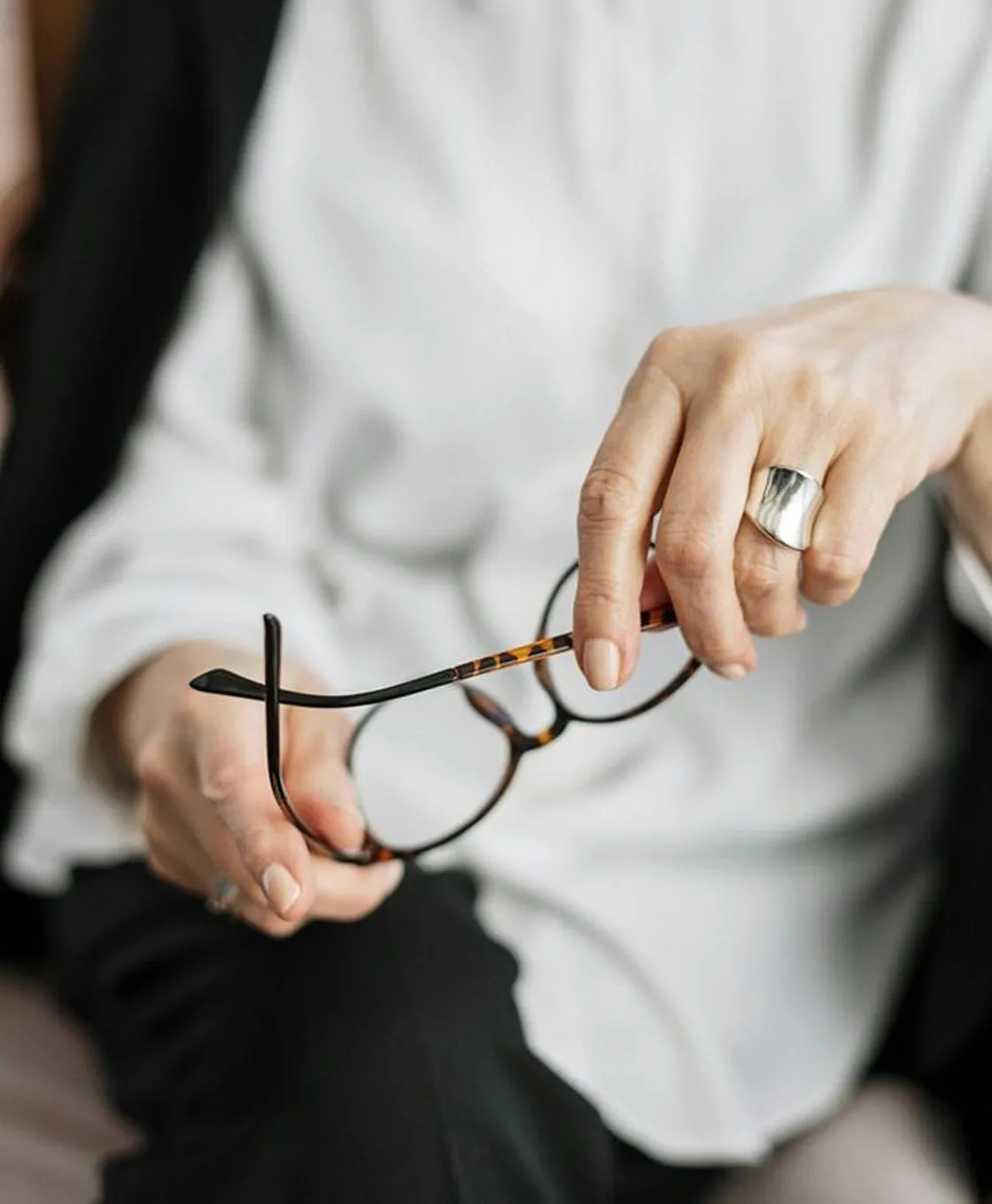 Close-up of a person holding tortoiseshell eyeglasses with both hands, wearing a silver ring and a white shirt.