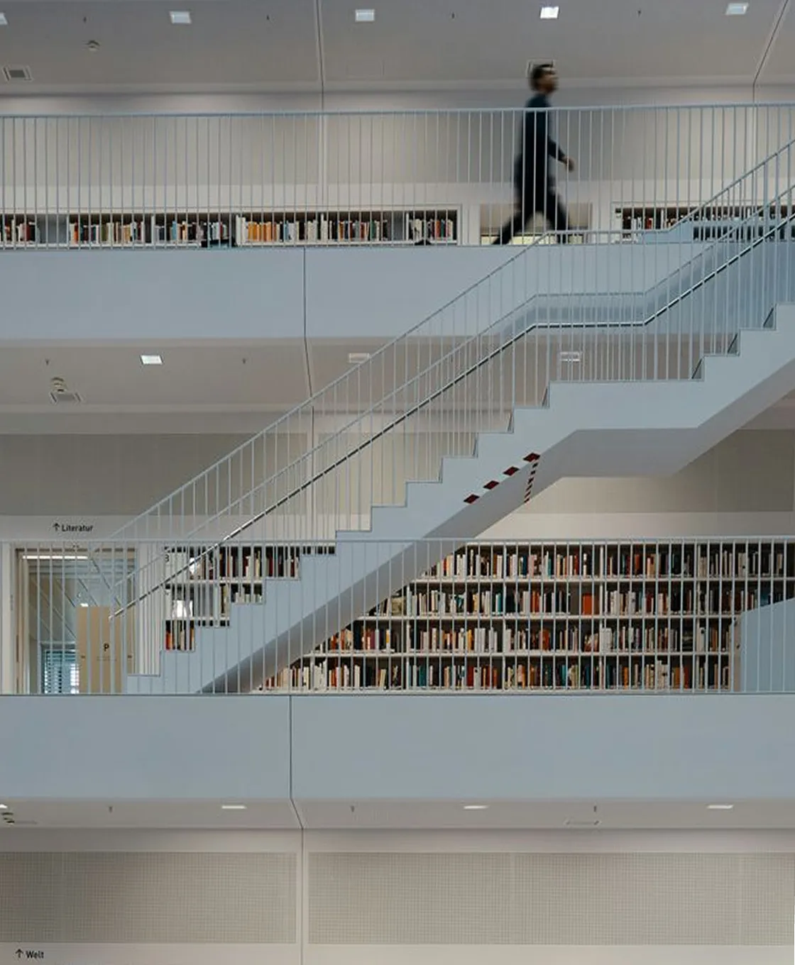 Modern library interior with white railings, a staircase, and rows of bookshelves filled with books, and a blurred person walking on the upper floor.