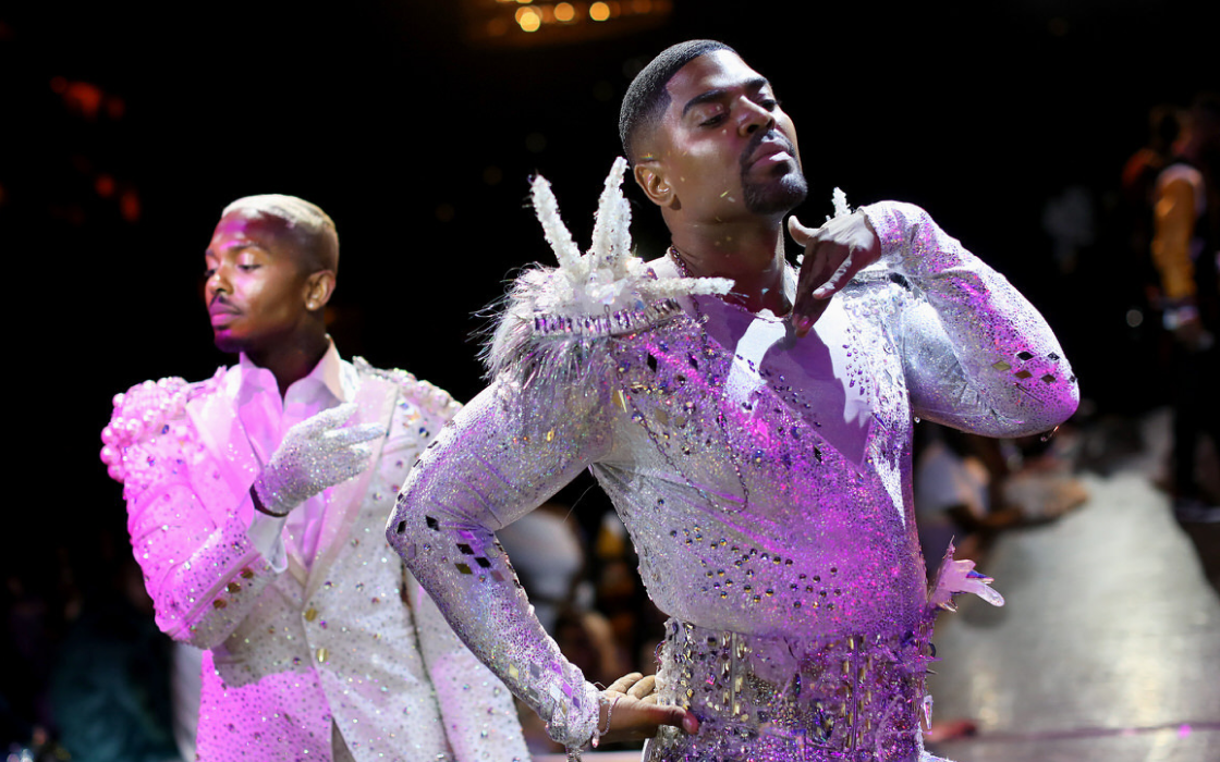 Two male dancers in glittering, embellished costumes performing on stage with dramatic lighting.
