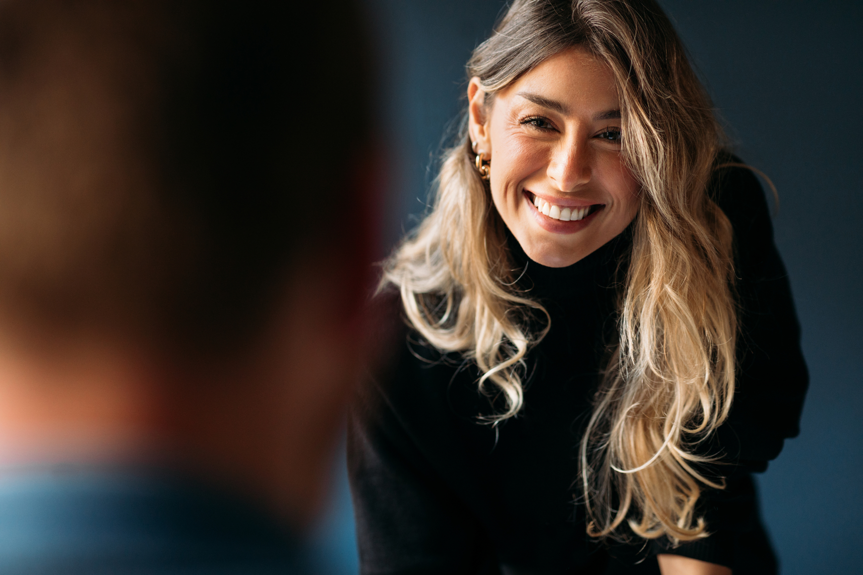 A woman with long blonde hair smiles  cheerfully at an out-of-focus person standing right in front of the camera lens