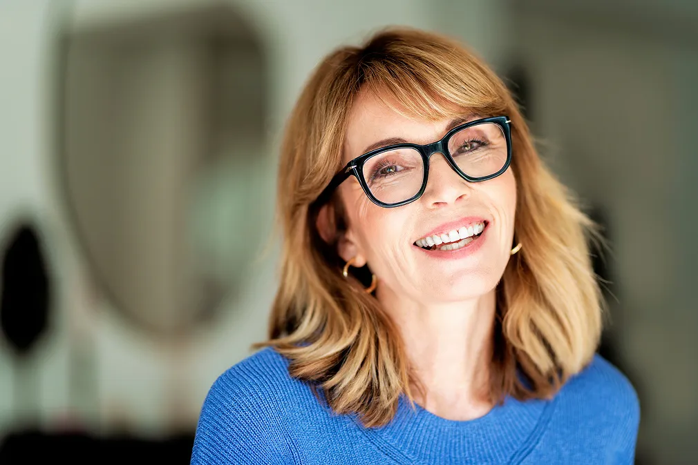 A woman with glasses smiling in front of a mirror.