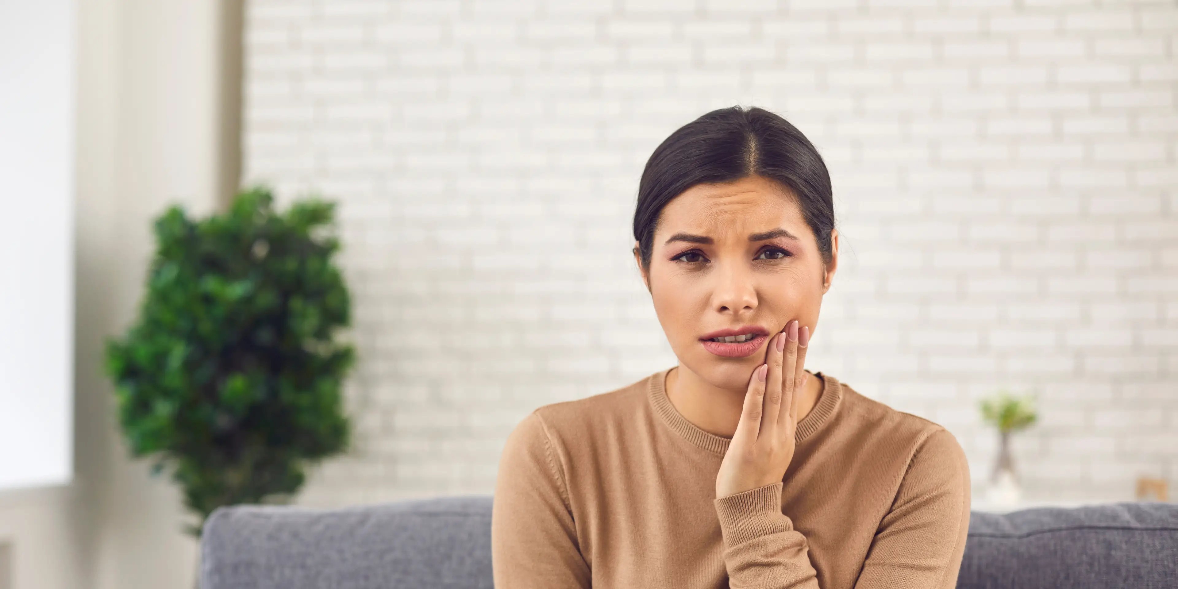 A woman sits on a couch, touching her cheek with a pained expression.