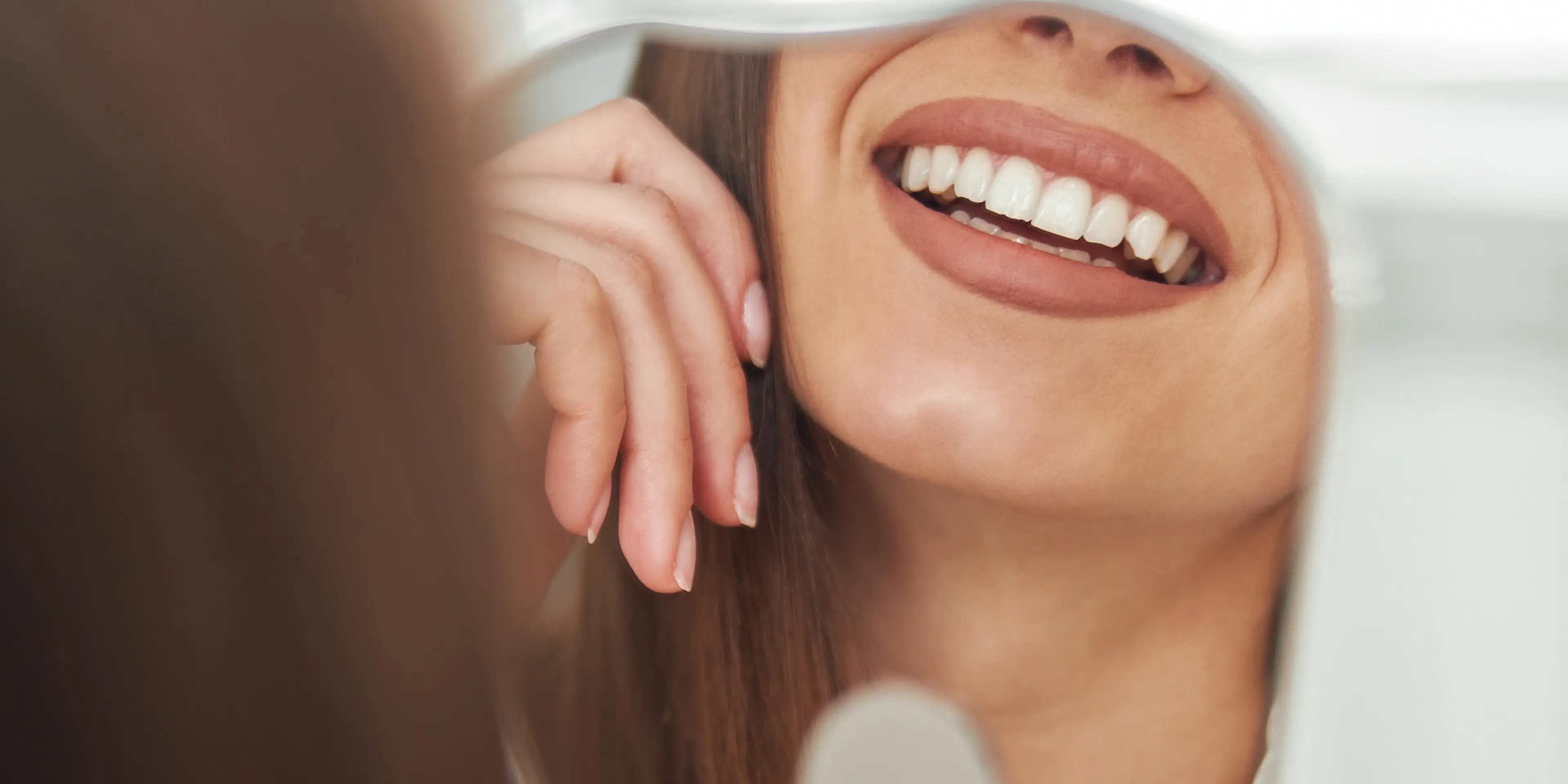 A person smiles brightly while looking at their teeth in a small mirror.