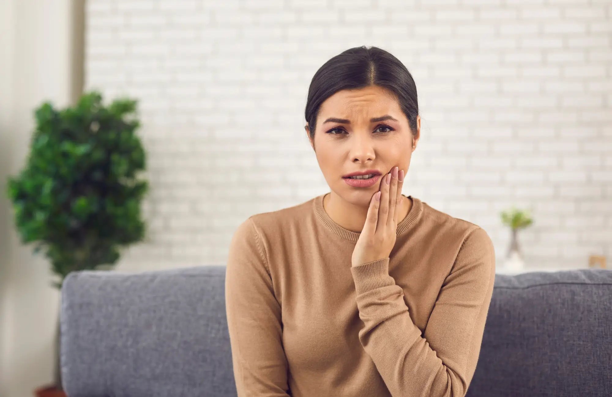A woman sits on a sofa, touching her cheek and looking concerned.
