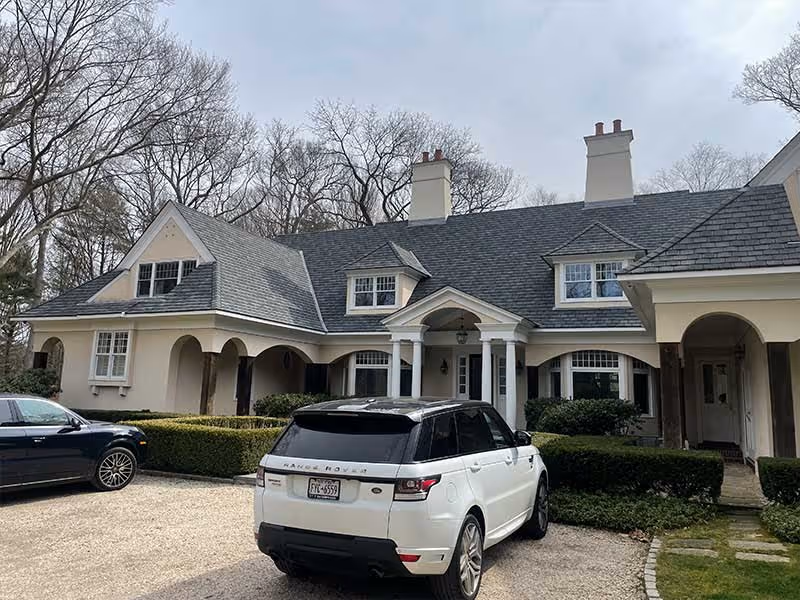 Large cream-colored house with gray shingle roof and two parked luxury SUVs in front.