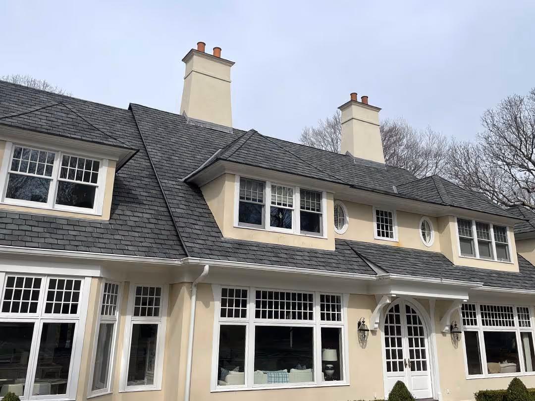 Large beige house with gray shingle roof, multiple white-framed windows, and two chimneys.