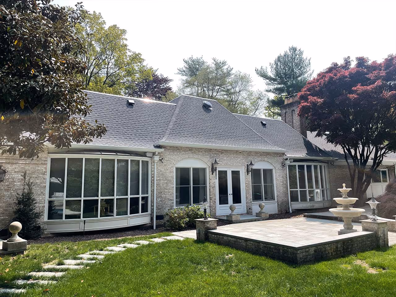 Backyard patio with stone tiles, a tiered water fountain, and a house with large windows and a shingle roof.