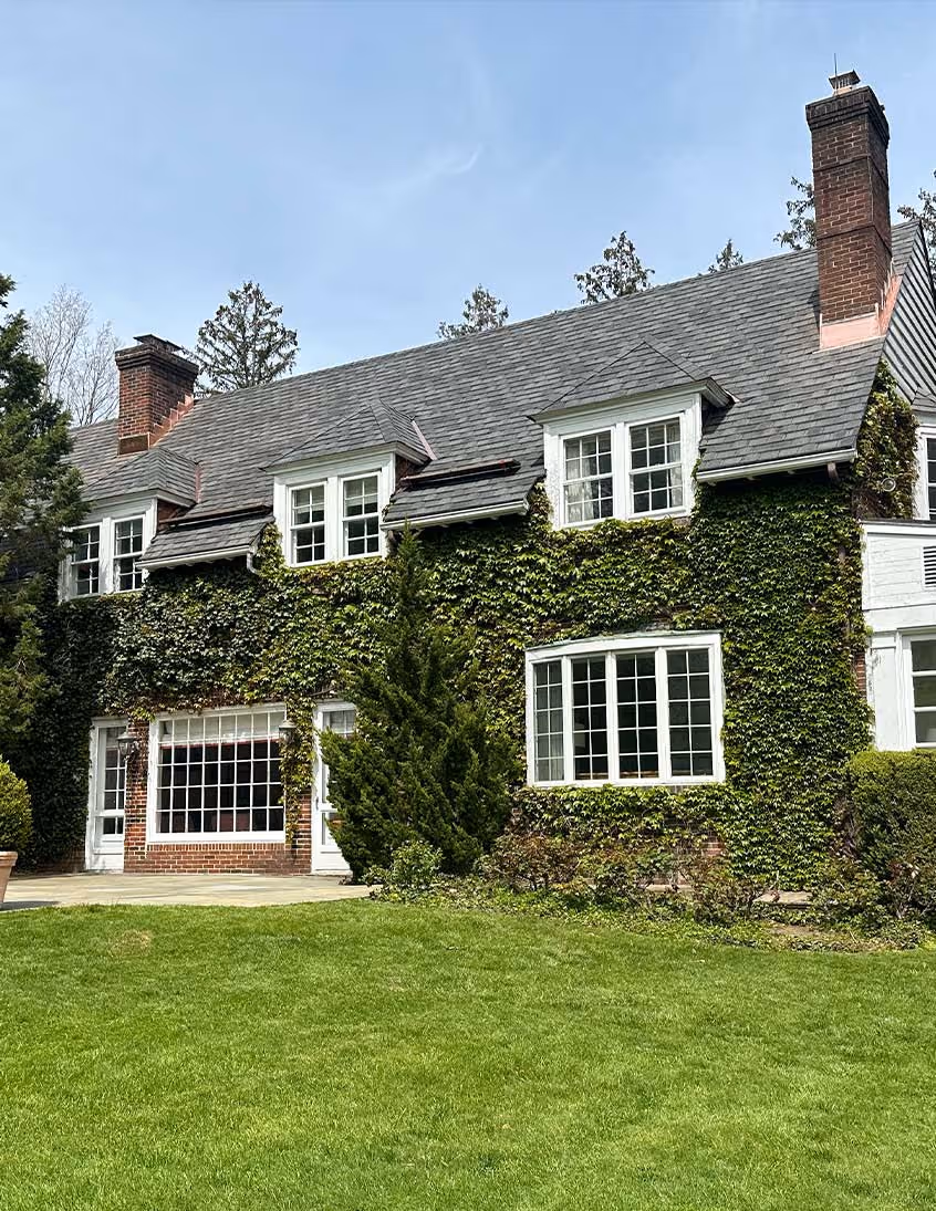 Two-story house covered with green ivy, featuring white-framed windows, a gray shingle roof, and two brick chimneys.