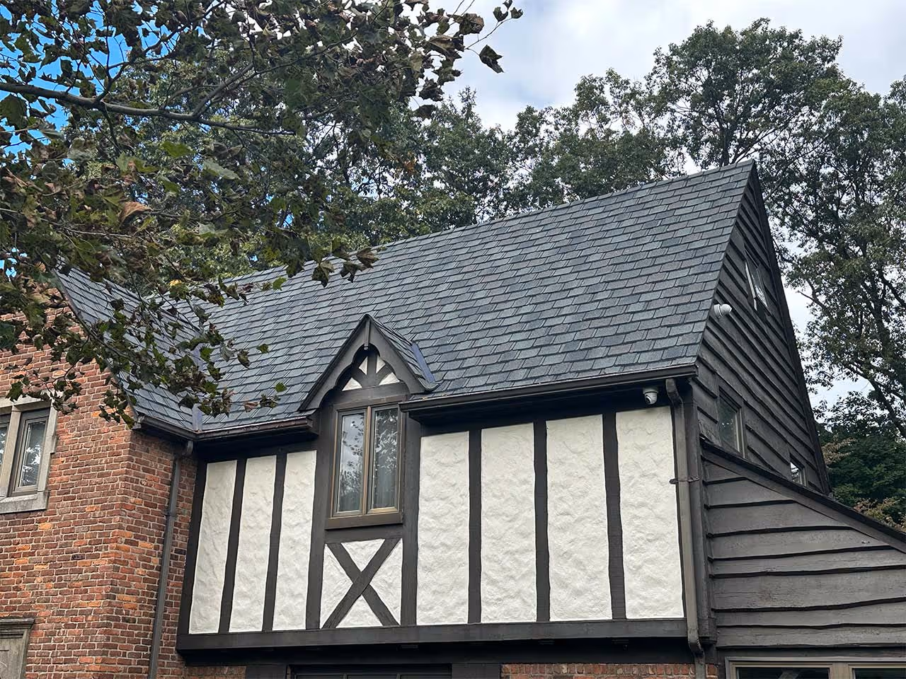 Tudor-style house section with gray shingle roof, timber framing, and brick wall with trees in background.