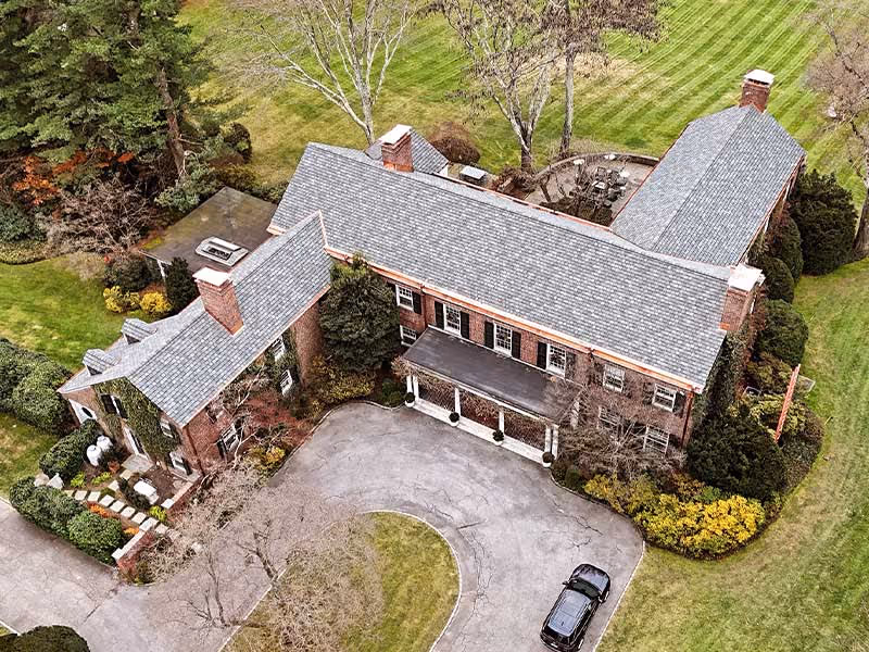 Aerial view of a large brick house with gray shingle roofs and surrounding green lawn.