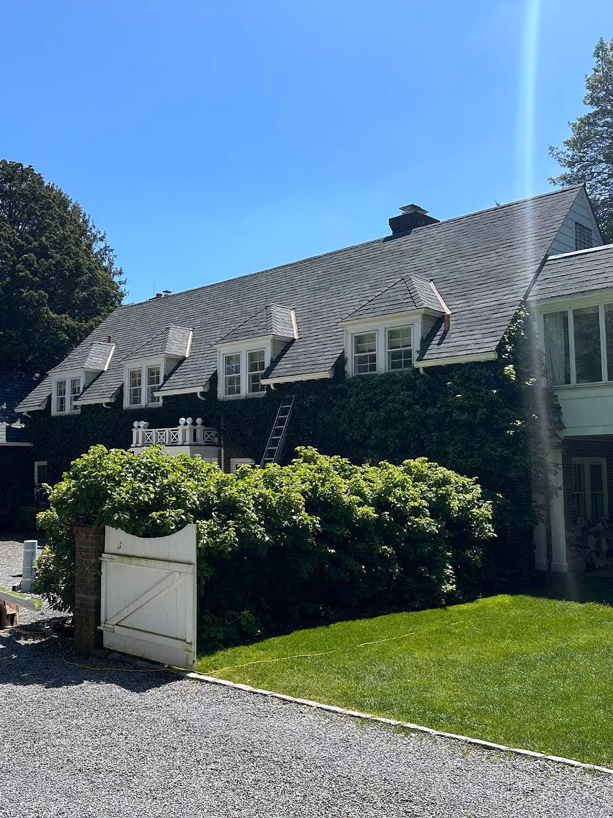 House with gray shingle roof, dormer windows, ivy-covered walls, green bushes, grass lawn, and a white wooden gate.