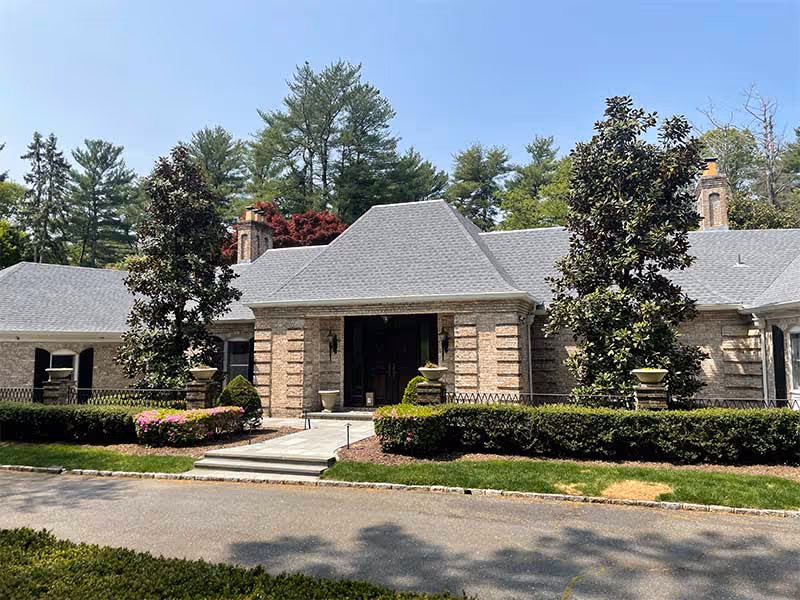 Large brick house with gray shingle roof, front entrance, landscaped bushes, and tall trees under clear blue sky.