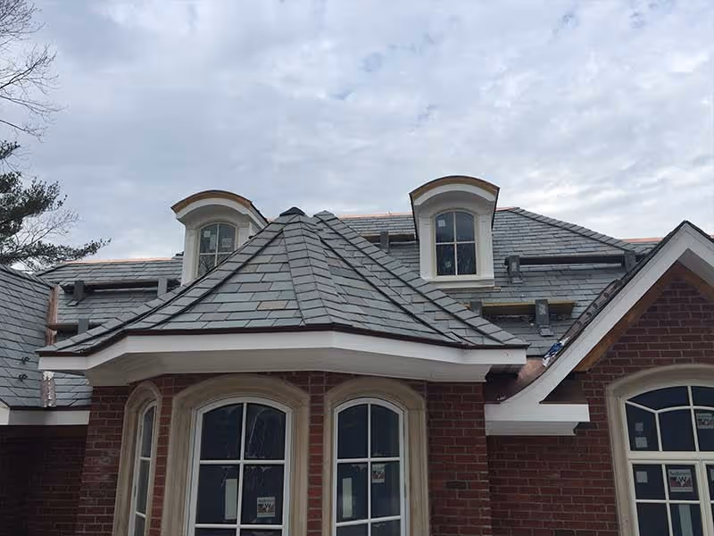 Brick house with multiple slate roof sections and dormer windows under a cloudy sky.