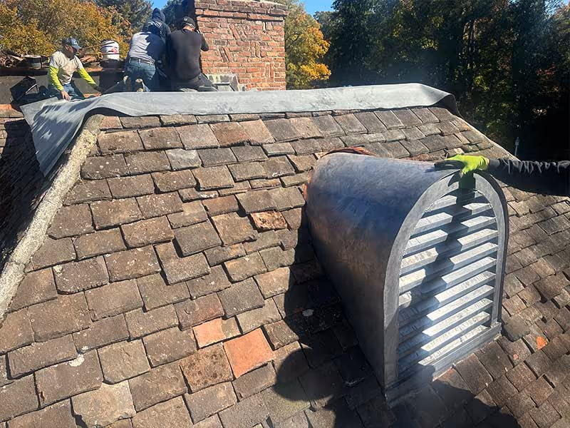 Three workers installing a metal vent on a weathered slate tile roof near a brick chimney in daylight.