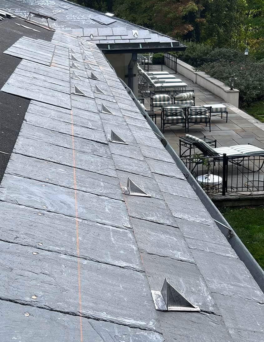 Slate roof installation with metal snow guards and terrace with loungers and chairs in the background.