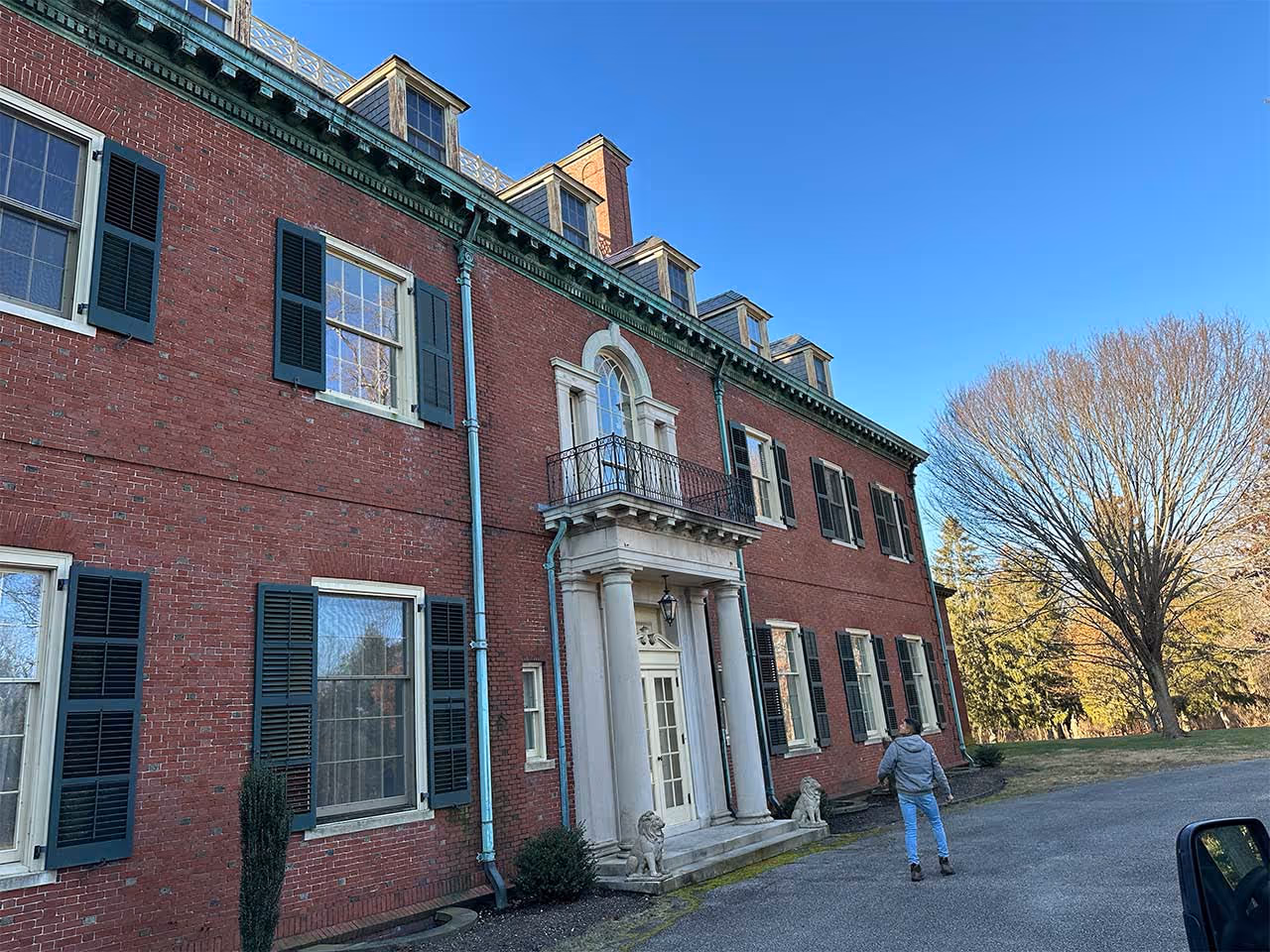 Person walking near a large red brick colonial-style building with green shutters and white columns.