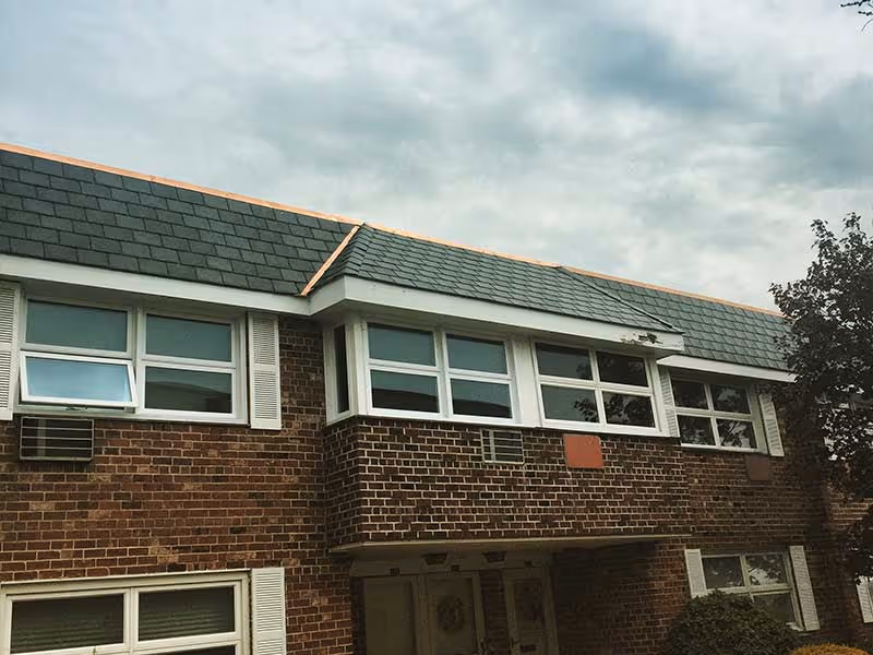 Brick house with slate roofing, multiple white-framed windows, and a cloudy sky.