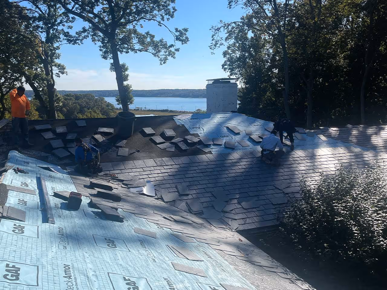 Workers installing slate roof shingles on a house with trees and a lake in the background.