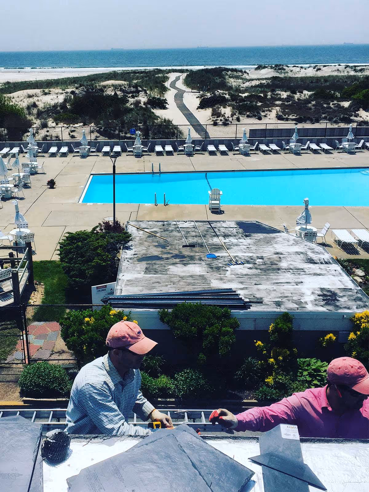 Two workers installing slate roofing on a building with a pool and sandy beach in the background.