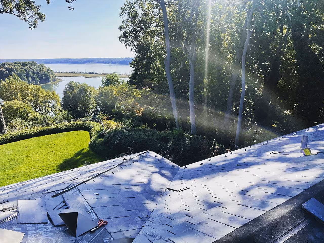 Rooftop under construction with slate tiles overlooking a green garden and distant water body.
