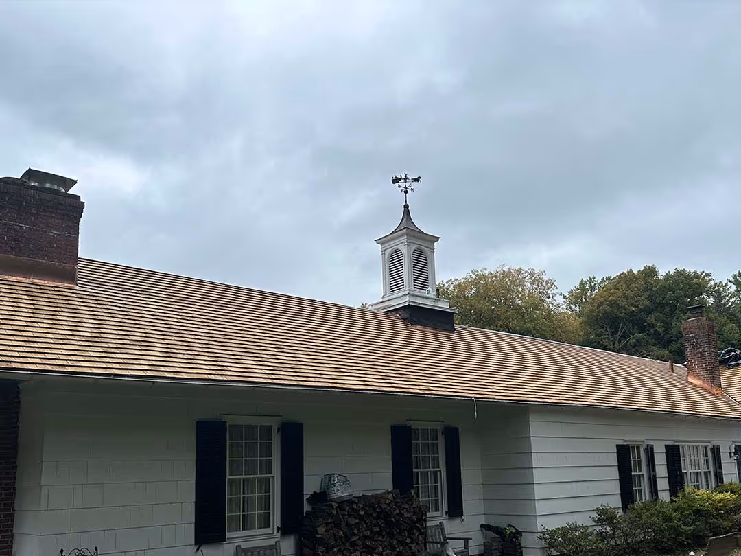 House with newly installed cedar shingle roof, white cupola, and weather vane under cloudy sky.